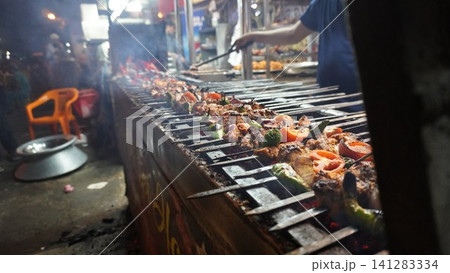 Long grill with kebab skewers cooking, smoke rising, outdoor setting at night, with dark shadowy areas. Long grill with kebab skewers cooking, smoke rising, outdoor setting at night, with dark shadowy areas. 141283334