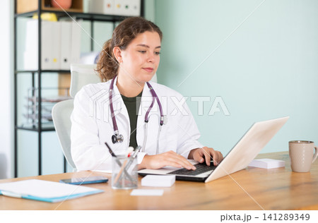 Young woman in white coat working with laptop in medical office Young woman in white coat working with laptop in medical office 141289349