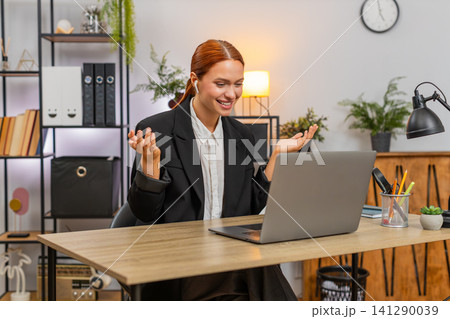 Young businesswoman joins video call on laptop wears earphones waves hi and gestures energetically 141290039