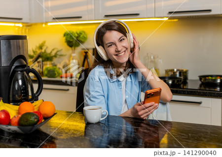 Young woman relaxing in home kitchen with headphones enjoying music while checking smartphone screen 141290040