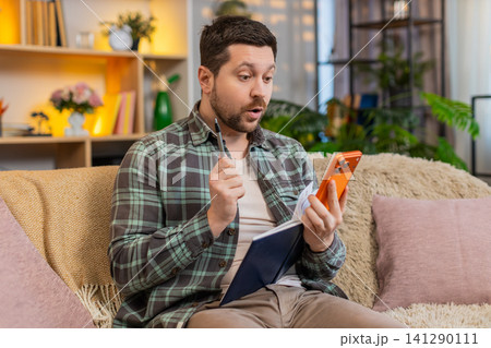 Young man on home sofa planning tasks with smartphone and notebook while checking messages at home Young man on home sofa planning tasks with smartphone and notebook while checking messages at home 141290111