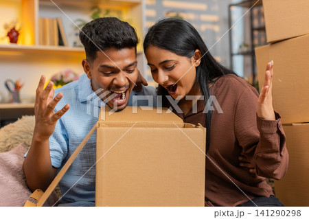 Indian couple man and woman sitting on home sofa opening delivery box with happy surprise reaction Indian couple man and woman sitting on home sofa opening delivery box with happy surprise reaction 141290298