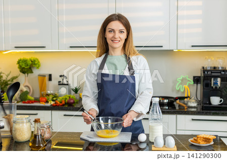 Middle-aged woman whisking eggs for omelet smiling in home kitchen for healthy diet breakfast recipe 141290300