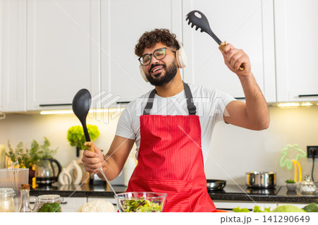 Arabian young adult man dancing in kitchen mixing fresh salad while listening to music in headphones Arabian young adult man dancing in kitchen mixing fresh salad while listening to music in headphones 141290649