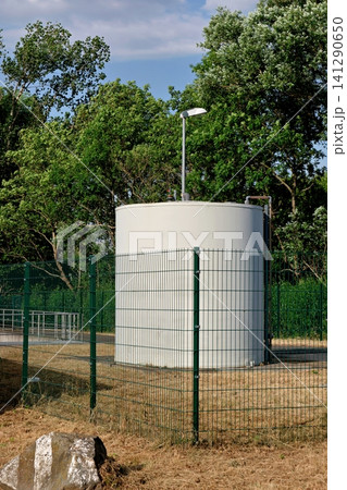 White industrial storage tank enclosed by green mesh fence with trees in background White industrial storage tank enclosed by green mesh fence with trees in background 141290650