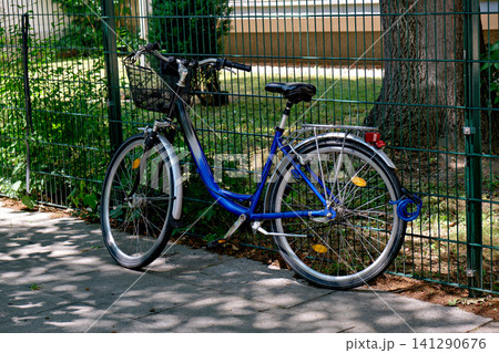 Blue city bicycle with basket parked next to green fence Blue city bicycle with basket parked next to green fence 141290676