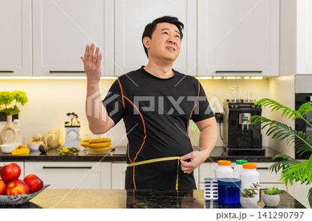 Heavy Asian adult man measures waist with tape upset about big belly fat weight gain at home kitchen Heavy Asian adult man measures waist with tape upset about big belly fat weight gain at home kitchen 141290797
