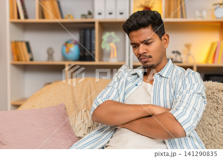 Indian man on home sofa frowning with dissatisfied look while sitting in thought in cozy living room 141290835