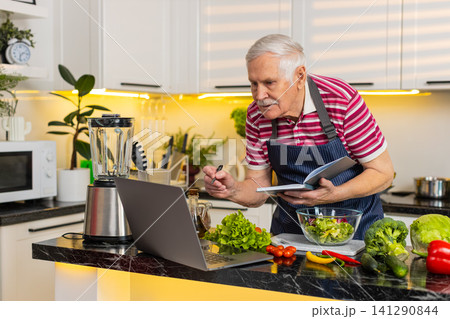 Senior old man making vegetable salad using laptop recipe and writing notebook notes for diet plan Senior old man making vegetable salad using laptop recipe and writing notebook notes for diet plan 141290844