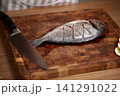 Fresh gilt-head bream fish with score marks, ready for cooking on a dark rustic wooden cutting board with a sharp knife 141291022