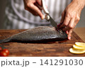 Chef's hands cleaning whole fresh sea bream with kitchen scissors on a rustic wooden cutting board for cooking 141291030
