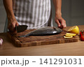 Chef's hands holding a knife, preparing a whole fresh sea bream on a wooden cutting board with lemon and tomatoes 141291031