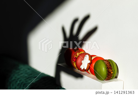 Halloween macarons with scary hand shadow on white wall. Line of colorful french macaroons with small ghost cookie on box, creative shadowplay, sweet food and autumn holiday concept Halloween macarons with scary hand shadow on white wall. Line of colorful french macaroons with small ghost cookie on box, creative shadowplay, sweet food and autumn holiday concept 141291363