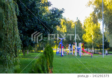 Empty colorful kids playground in green public park surrounded by trees. Beautiful summer landscape with children play area, slides, swings and green lawn, urban recreation and family leisure concept Empty colorful kids playground in green public park surrounded by trees. Beautiful summer landscape with children play area, slides, swings and green lawn, urban recreation and family leisure concept 141291373
