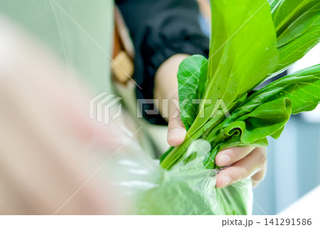 Closeup of people shopping for fresh green leafy vegetables and carrots at a local market stall, with hands selecting produce for purchase Closeup of people shopping for fresh green leafy vegetables and carrots at a local market stall, with hands selecting produce for purchase 141291586