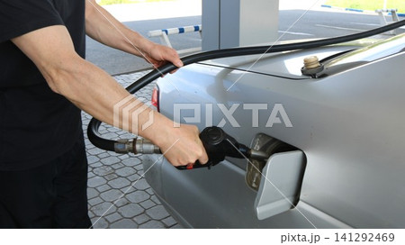 A man in black filling up a silver car at a gas station. He holds the fuel nozzle inserted into the gas tank door and holds a long black hose with his hand. The man is filling up a car at a gas pump. A man in black filling up a silver car at a gas station. He holds the fuel nozzle inserted into the gas tank door and holds a long black hose with his hand. The man is filling up a car at a gas pump. 141292469