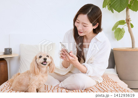 ベッドの上で愛犬と一緒にスマートフォンを操作する笑顔の若い女性 ベッドの上で愛犬と一緒にスマートフォンを操作する笑顔の若い女性 141293619