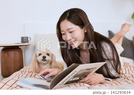 ベッドに寝転びながら愛犬と一緒に雑誌を読む笑顔の若い女性 141293634