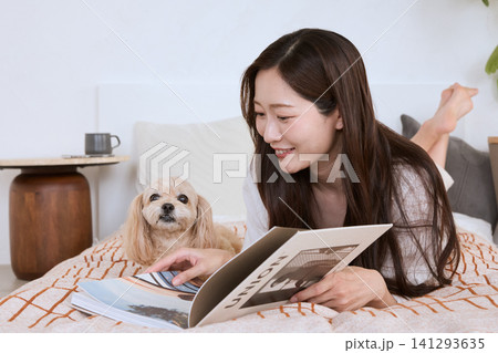 ベッドに寝転びながら愛犬と一緒に雑誌を読む笑顔の若い女性 ベッドに寝転びながら愛犬と一緒に雑誌を読む笑顔の若い女性 141293635