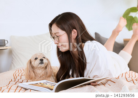 ベッドに寝転んで愛犬と一緒に本を読む眼鏡の女性 ベッドに寝転んで愛犬と一緒に本を読む眼鏡の女性 141293656