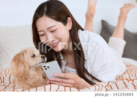 ベッドに寝転びスマートフォンを見る女性と愛犬 ベッドに寝転びスマートフォンを見る女性と愛犬 141293693