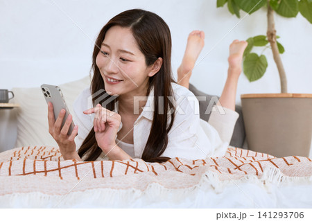 ソファでくつろぎながらスマートフォンを操作する笑顔の若い女性 ソファでくつろぎながらスマートフォンを操作する笑顔の若い女性 141293706