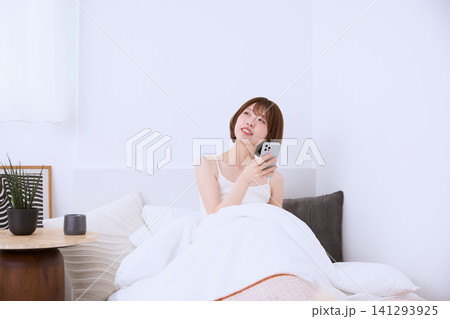 ベッドの上でスマートフォンを持ち微笑む若い女性 ベッドの上でスマートフォンを持ち微笑む若い女性 141293925