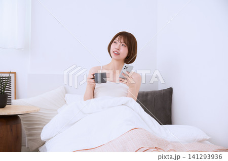 ベッドの上でスマートフォンとコーヒーを持つ若い女性の朝の時間 ベッドの上でスマートフォンとコーヒーを持つ若い女性の朝の時間 141293936