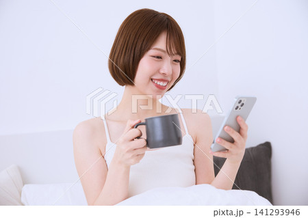 ベッドの上でスマートフォンを見ながらコーヒーを飲む笑顔の若い女性 141293946