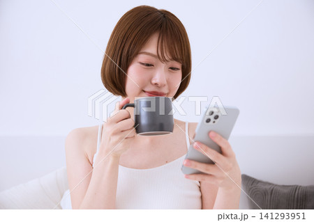 ソファでスマートフォンを見ながらコーヒーを飲む若い女性 ソファでスマートフォンを見ながらコーヒーを飲む若い女性 141293951