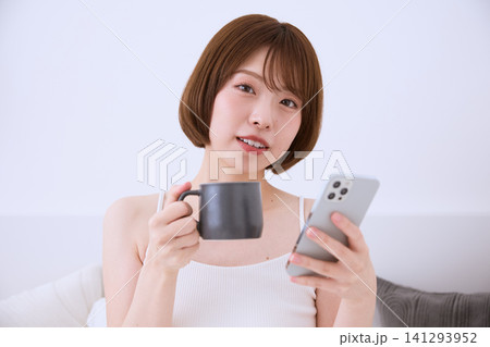 スマートフォンとコーヒーカップを持ちくつろぐ若い女性 スマートフォンとコーヒーカップを持ちくつろぐ若い女性 141293952