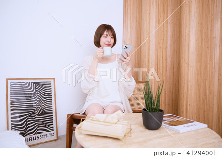 スマートフォンを見ながらコーヒーを飲むリラックスした若い女性 スマートフォンを見ながらコーヒーを飲むリラックスした若い女性 141294001