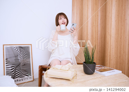 自宅でスマートフォンを見ながらコーヒーを飲むリラックスした若い女性 自宅でスマートフォンを見ながらコーヒーを飲むリラックスした若い女性 141294002
