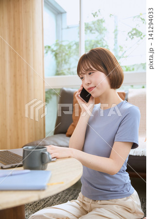 リビングでスマートフォンで電話しながらノートパソコンを操作する若い女性 141294033