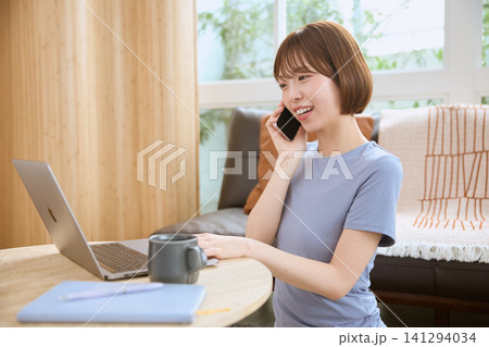 リビングでスマートフォンで通話しながらノートパソコンを使う若い女性 141294034
