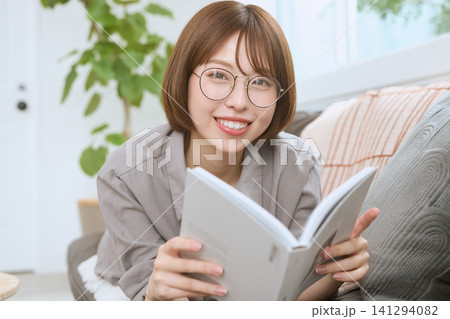 ソファで本を読むメガネをかけた笑顔の若い女性 141294082