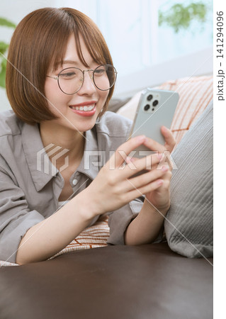 ソファでスマートフォンを見て笑顔の眼鏡をかけた若い女性 ソファでスマートフォンを見て笑顔の眼鏡をかけた若い女性 141294096