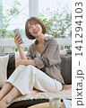 ソファに座りスマートフォンを見て笑顔の若い女性 141294103