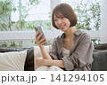 ソファに座りスマートフォンを見て笑顔の若い女性 141294105