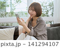ソファに座りスマートフォンを見て微笑む若い女性 141294107