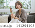 ソファに座りスマートフォンを持つ笑顔のボブヘアの若い女性 141294108