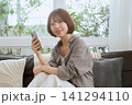 リビングのソファでスマートフォンを持ち微笑む若い女性 141294110