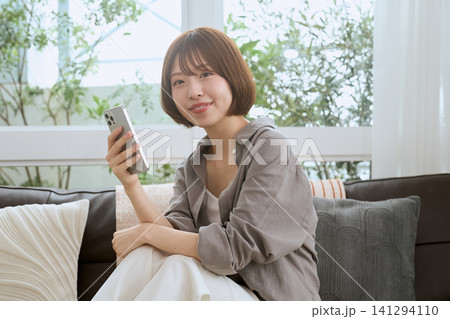 リビングのソファでスマートフォンを持ち微笑む若い女性 リビングのソファでスマートフォンを持ち微笑む若い女性 141294110