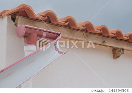 Horizontal close-up view of a broken plastic roof gutter detached and hanging from a residential house wall under a red clay tile roof during bad weather Horizontal close-up view of a broken plastic roof gutter detached and hanging from a residential house wall under a red clay tile roof during bad weather 141294508