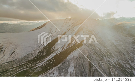 Horizontal aerial view of magnificent Caucasian mountain peaks covered with light snow under heavy dramatic clouds illuminated by warm sunset light in Georgia Horizontal aerial view of magnificent Caucasian mountain peaks covered with light snow under heavy dramatic clouds illuminated by warm sunset light in Georgia 141294558