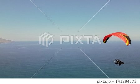 Horizontal scenic view of a paratrike with a red and yellow wing flying high over the calm blue sea water near coast on a sunny day for summer holiday promo 141294573