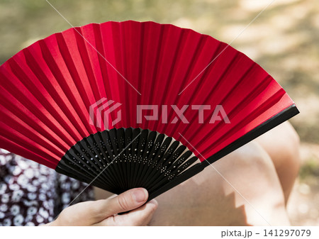 Woman in a floral patterned dress holding a red folding fan in a courtyard, illustrating heatwave lifestyle adaptations in Europe. Woman in a floral patterned dress holding a red folding fan in a courtyard, illustrating heatwave lifestyle adaptations in Europe. 141297079