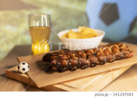 Grilled ribs on a wooden board, beer and snacks arranged for a soccer viewing party, creating a cozy football night atmosphere. Grilled ribs on a wooden board, beer and snacks arranged for a soccer viewing party, creating a cozy football night atmosphere. 141297083
