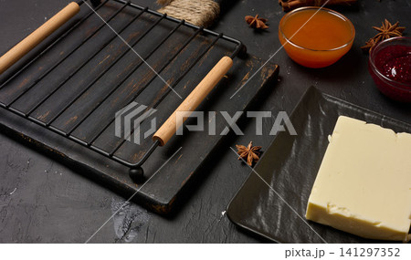 Kitchen still life with cutting board, baking rack, butter and jam on dark background, top view 141297352