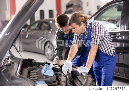 Two mechanics examining car engine under open hood in workshop Two mechanics examining car engine under open hood in workshop 141297713